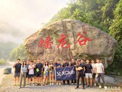 A group of people poses for a photo in front of a large rock inscribed with the red Chinese characters "蟠龙谷," while one person holds a blue "XClub" banner.