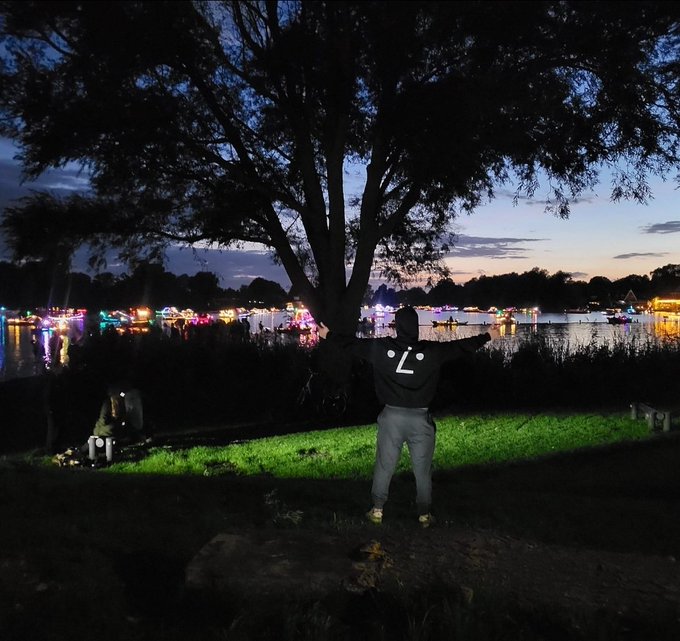 A silhouetted figure wearing a hoodie with a smiley face logo stands with their back to the camera, looking out at a lit-up lakeside festival at dusk.