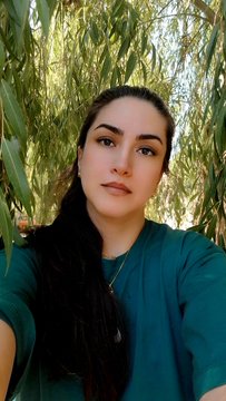 A young woman with long dark hair takes a selfie outdoors, standing under a tree with green leaves, wearing a teal shirt and a delicate necklace.