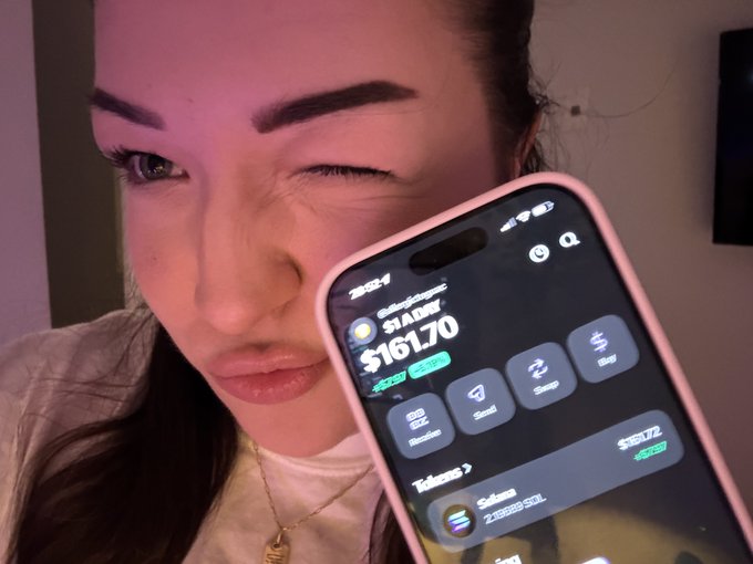 In the image, a woman with brown hair is holding her cell phone up to her face. She appears to be making a funny face for the camera while showing off the contents of her smart device screen. The phone's display features several app icons and information related to online shopping activities, including an app named "SOL". One particular message on the screen reads "day 213 of buying $1 worth of SOL until ATH WE GOIN UPPP", which could indicate that she is checking out a new store or product during a specific day.
