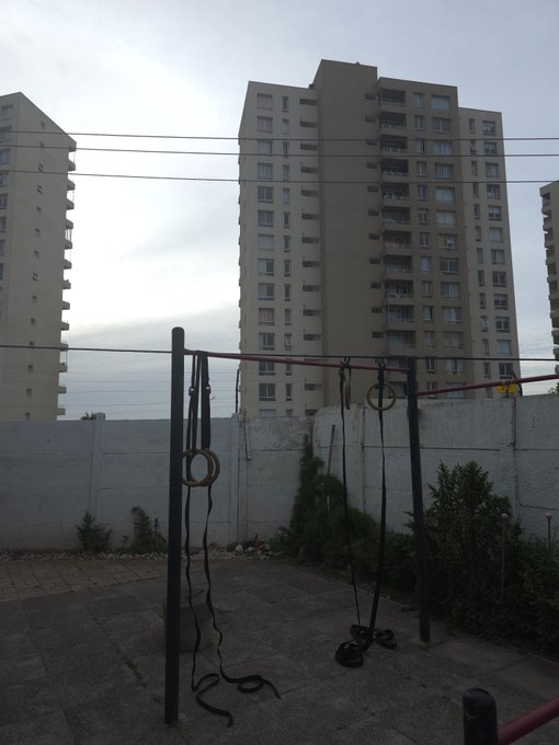 An outdoor calisthenics gym with pull-up bars, gymnastic rings, and resistance bands set up on a concrete pad, with a tall beige apartment building in the background.