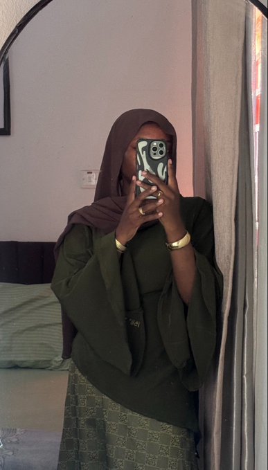 A mirror selfie of a woman wearing a dark brown hijab and green outfit, holding a phone that covers her face.