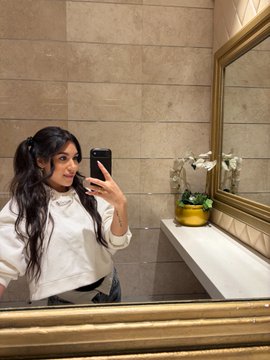 A mirror selfie of a woman with long dark hair taking a photo with her phone in what appears to be an upscale bathroom with marble walls, a gold-framed mirror, and a small potted orchid on the counter.