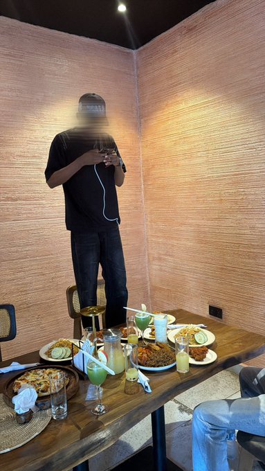 A person in a black shirt and jeans stands holding a phone with a white charging cable, next to a wooden dining table filled with plates of food, drinks, and a woven placemat in a warmly lit room.
