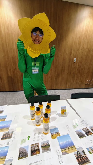The image features a person dressed up as a flower, wearing a green outfit with yellow paper flowers on their head. They are standing at a table filled with various items such as bottles of oil paint and pictures. The person appears to be posing for the photo while holding one of these items in front of them.