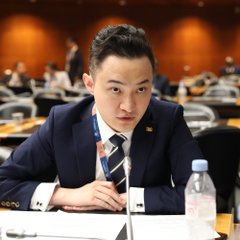 A man in a dark suit and blue tie sits at a conference table with a water bottle and documents, wearing a "TGE20" badge on his lanyard. The image likely relates to the post as the individual pictured may be Justin Sun, founder of Tron, given the formal conference setting consistent with crypto industry events where such quantum cryptography announcements would be made.