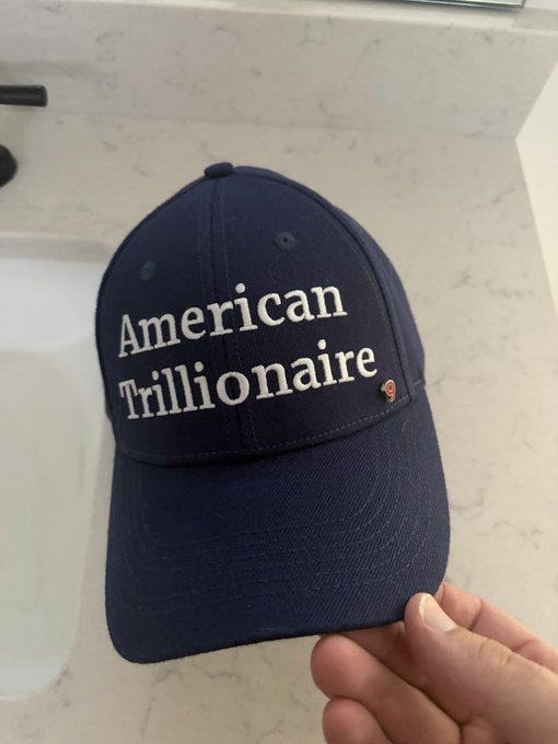 A navy blue baseball cap is held up against a marble countertop, featuring white embroidered text that reads "American Trillionaire" in a classic serif font. The cap serves as a fitting patriotic accessory for the 4th of July, as referenced in the caption celebrating a notable piece from gmoneyNFT.