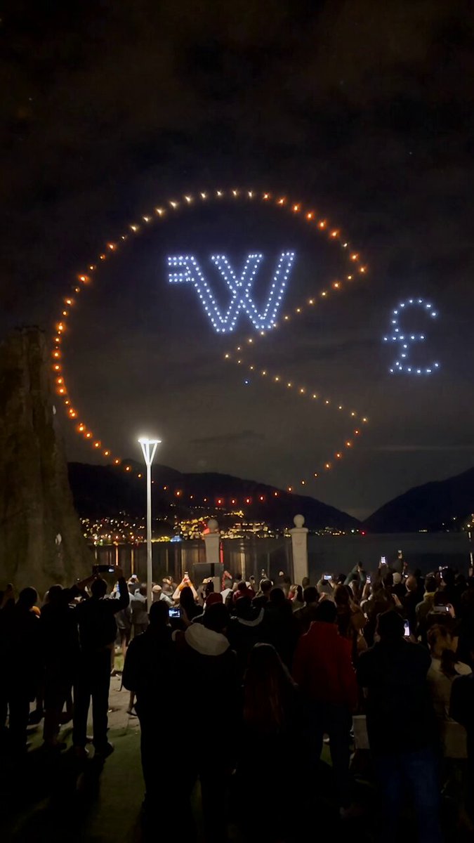 The image shows a large crowd of people gathered around an event, with a giant billboard featuring the letters "W" and "E" flying through the air. The billboard displays the text "Giant Bitcoin drone show in Switzerland showing $WORTHLESS eating fiat currencies like Pac-Man". This suggests that there is some sort of Bitcoin-related event or promotion taking place at this location, possibly involving the use of drones to display the Bitcoin symbol and its value.

The crowd appears to be engaged and interested in the event, as they are standing close together and looking up towards the billboard. The presence of a large number of people indicates that it is an important occasion or attraction for those who have gathered around to see what's happening.