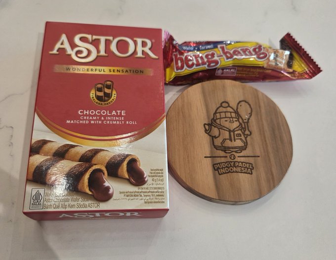 A box of Astor chocolate wafer sticks and a Bing-bang candy bar sit next to a round wooden coaster engraved with 'PUDGÝ DADEL INDONESIA' and a cartoon character.