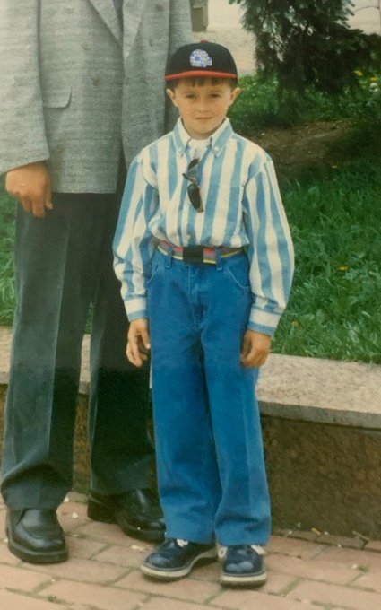 A vintage photograph of a young boy wearing a striped blue and white shirt, blue jeans, a baseball cap, and sunglasses hanging from his shirt, standing next to an adult in a gray suit.