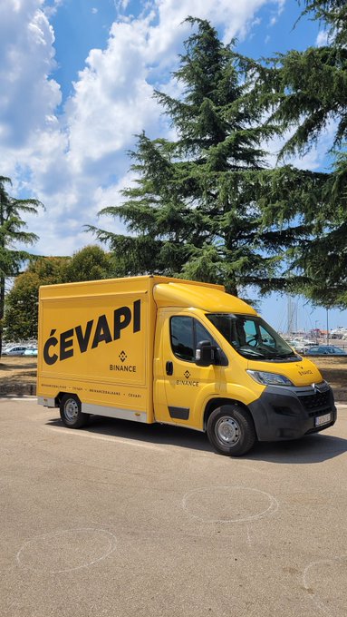 A yellow delivery truck branded with 'ĆEVAPI' and 'BINANCE' logos, parked in a lot with large trees and a blue sky in the background.