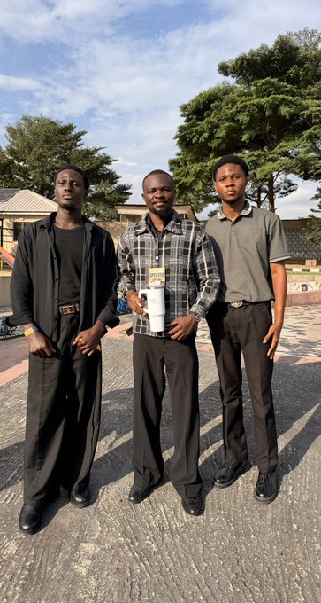 Three young men pose together outdoors at the Lagos web3 hangout event, with the man in the center wearing a plaid shirt and a lanyard, flanked by his companions in dark attire against a backdrop of trees and a partly cloudy sky.