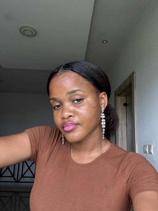 A selfie of a young woman with dark hair pulled back, wearing a brown ribbed shirt and long gold earrings, standing indoors near a staircase.