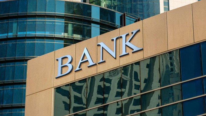 The image shows a building facade with large metallic letters spelling "BANK" mounted on a tan stone surface, set against a backdrop of reflective blue glass windows. This relates to the post about small U.S. banks opposing crypto companies receiving banking licenses, illustrating the banking industry context of the lawsuit filed by the Independent Community Bankers of America (ICBA) against the OCC.