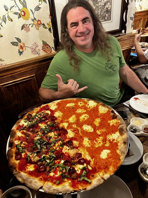 A man with long brown hair wearing a green t-shirt smiles and gestures at a large, mostly plain cheese pizza with one half topped with vegetables and olives, sitting at a restaurant table.