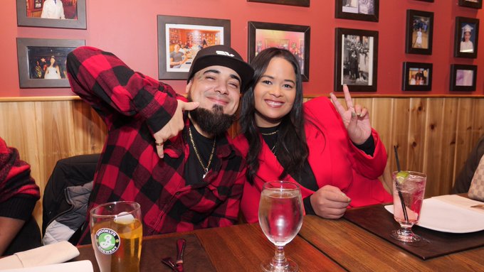 A man in a red plaid shirt and black cap and a woman in a red sweater sit together at a restaurant table, smiling and holding up peace signs next to a beer glass.