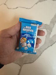 A hand holds up a small blue packet of 'Rice Krispies' candy on a tiled floor.