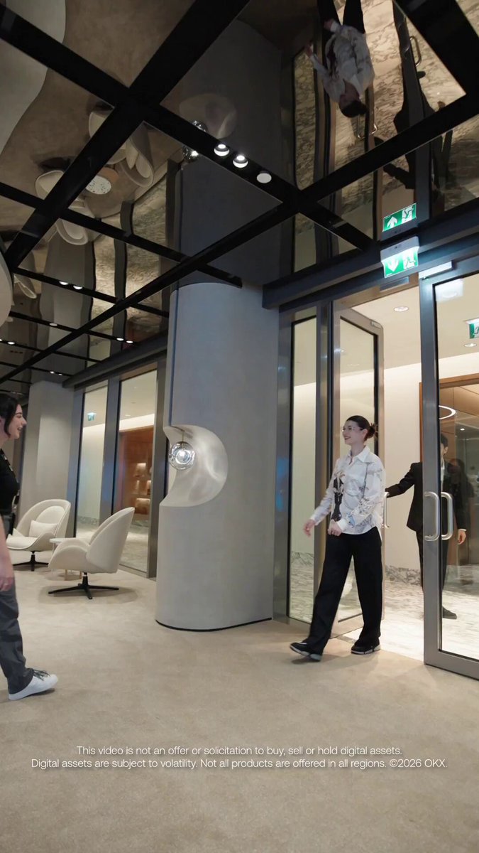 The image shows a group of people standing inside an office building with large windows that reflect the surroundings outside, including a person holding a camera. The room has a modern design featuring a glass ceiling and white walls. A man wearing a tie can be seen in the background, possibly attending to some work-related tasks or taking part in a meeting. The image also contains text about an event happening on June 11th at OKX, which seems to be related to the office space where people are gathered.