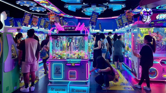 A crowded, neon-lit arcade filled with players gathered around glowing claw machines and prize games, with "SUCKYOO" branding and "MIO AV" signage visible throughout the vibrant venue.

The image visually echoes the post's message by depicting real people physically engaging with arcade claw machines—the same machines referenced as an everyday example of revenue-generating assets—while $PLAY from DualMintRWA represents the onchain innovation making such real-world yields accessible and verifiable.