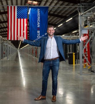Eric Trump stands with arms outstretched in a warehouse, with an American flag and a blue banner reading "AMERICAN B" visible behind him, celebrating American Bitcoin's milestone of surpassing 8,000 BTC after a 500 BTC addition.