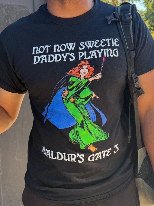 A person wears a black t-shirt featuring a comic-style red-haired witch holding a staff, with text reading 'NOT NOW SWEETIE DADDY'S PLAYING' above and 'BALDUR'S GATE 3' below.