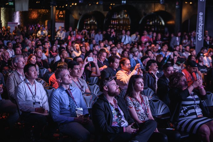 A large, attentive audience sits in rows at a dimly lit event with a 'coinsquare' banner visible on the right side.