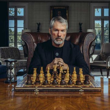 A man with grey hair and a beard sits behind a wooden chessboard in a classic room, wearing a black collared shirt and a necklace, with an hourglass visible on the left.