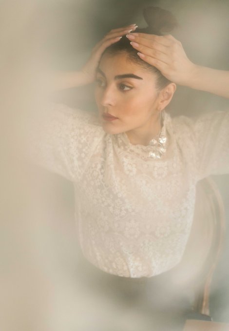 A woman wearing a white lace top with her hands resting on her head, photographed in soft, hazy lighting.