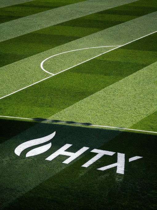 A close-up view of a green striped soccer field displaying the Al Hilil Saudi FC logo painted in white on the grass.