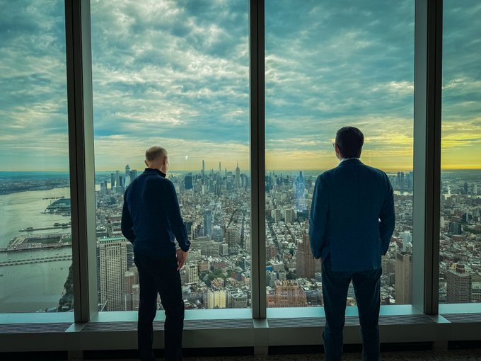 Two men in suits stand with their backs to the camera, looking out through floor-to-ceiling windows at a sprawling city skyline at sunset.