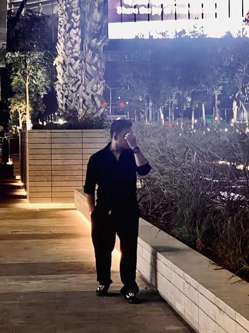 A nighttime photo of a man dressed in black standing on a paved walkway, holding his hand up to his face, with illuminated trees and streetlights in the background.
