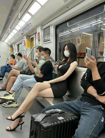 Passengers seated inside a subway or train car, several looking at their phones; a woman in a black dress and heels sits next to a black suitcase.