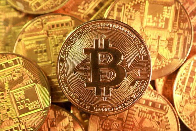 A close-up photograph of multiple gold-colored physical Bitcoin tokens stacked together, with the prominent 'B' symbol visible on the central coin.