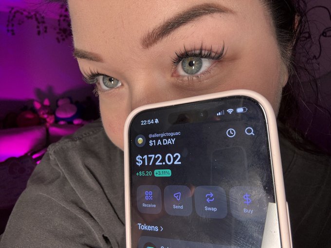 A close-up selfie of a young woman with blue eyes partially hidden behind her phone, which displays a crypto wallet showing $172.02 (+$5.20, +3.11%) for the account @allergictoguoac.