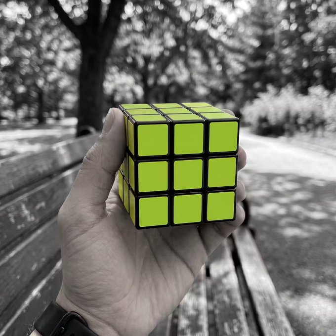 A hand holds a solved Rubik's Cube, with the bright yellow face standing out against the otherwise black-and-white park background featuring a wooden bench and trees. The vibrant yellow echoes the "GM" (good morning) post, as the cube's cheerful color mirrors the upbeat morning greeting.