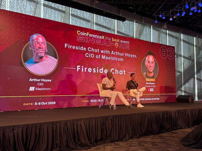Two men sit on stage during a 'CoinFerenceX Singapore' event, participating in a 'Fireside Chat' with Arthur Hayes, CIO of Maelstrom, as displayed on the large screen behind them.