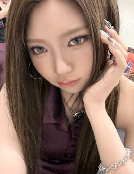 A close-up portrait of a young woman with long brown hair, wearing a dark purple collared top, a necklace, and large rings, posing with her hand near her face.