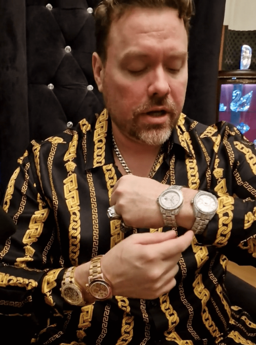 A man in a black and gold Versace-style patterned shirt, displaying multiple luxury watches on both wrists and a ring.