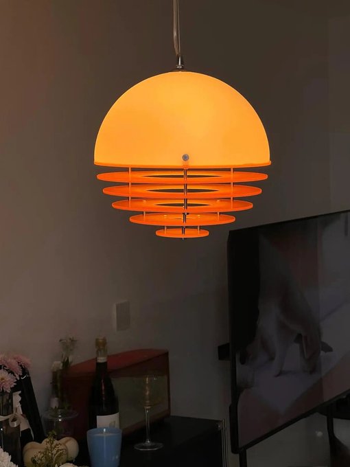 A warm orange pendant lamp with a dome-shaped top and tiered circular shades glows in a dimly lit room, with a wine bottle, flowers, and a television visible below.