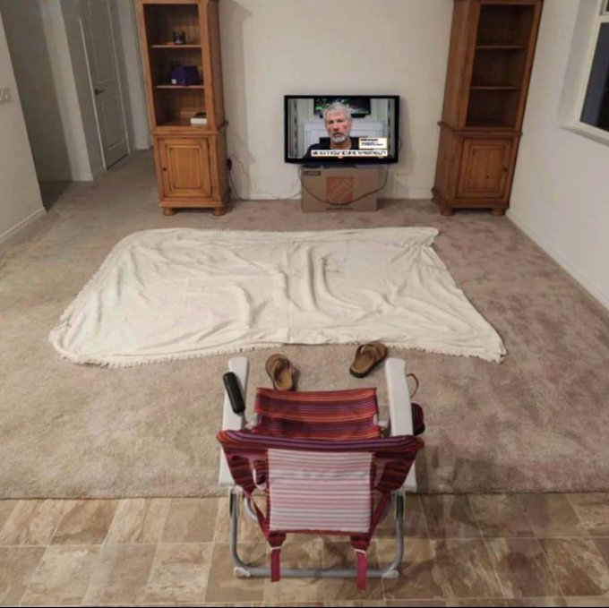 A living room setup with a beach chair facing a TV displaying a man's face, with a blanket spread out on the carpet between them like a makeshift outdoor viewing area.
