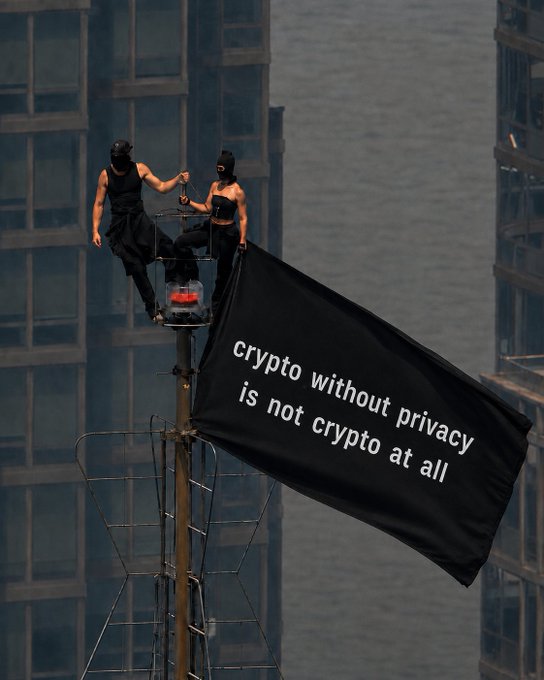 Two masked individuals stand atop a structure beside a large black flag, set against an urban skyline. The flag reads, "crypto without privacy is not crypto at all," reinforcing the post's message that privacy is essential to the true meaning of cryptocurrency.