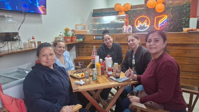 Five women are seated around a wooden table in a restaurant with a neon 'M' sign and orange balloons in the background, with condiments and drinks on the table.