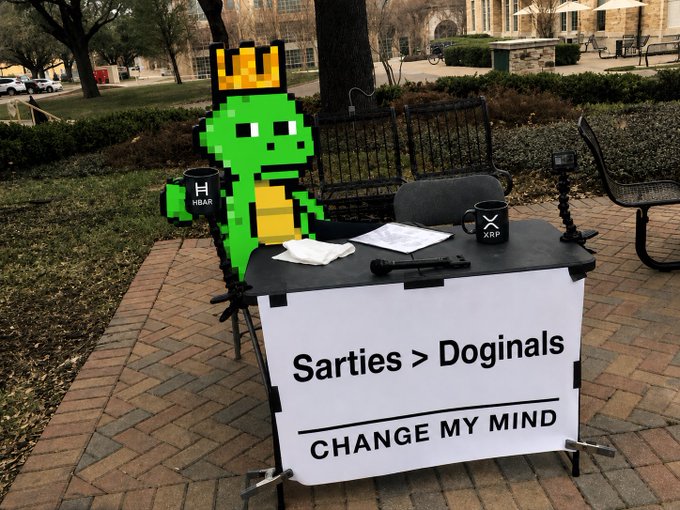 A pixelated green dinosaur wearing a crown sits behind a table featuring XRP and HBAR mugs, alongside a sign reading "Sarties > Doginals - CHANGE MY MIND," playfully challenging viewers to argue that Doginals are superior to Sartoshi NFT collections.