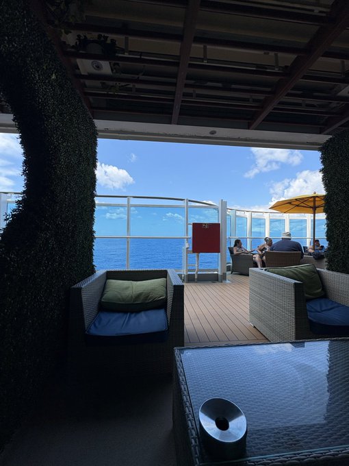 A shaded outdoor lounge area on a cruise ship deck featuring wicker seating with cushions, a small pool or fountain in the foreground, and an ocean view with passengers relaxing under an umbrella.