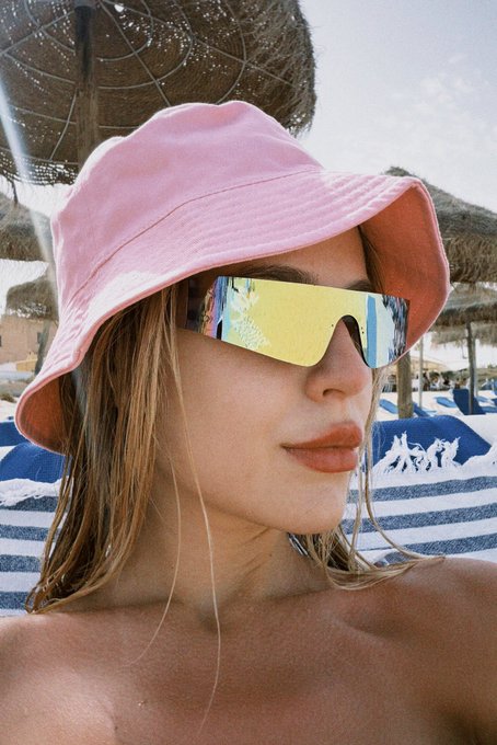 A woman with wet hair wearing a pink bucket hat and reflective yellow sunglasses, posing at a beach resort.