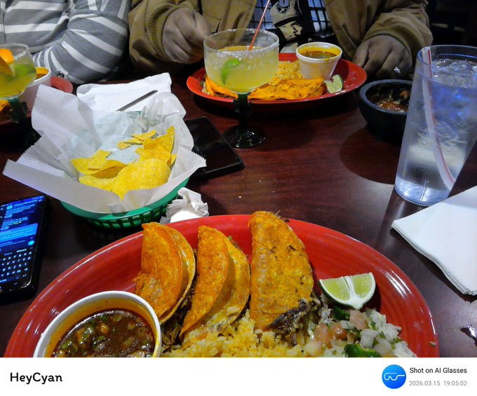 A restaurant meal featuring tacos on a red plate with rice, beans, salsa, and tortilla chips, accompanied by margaritas on a dark wooden table.