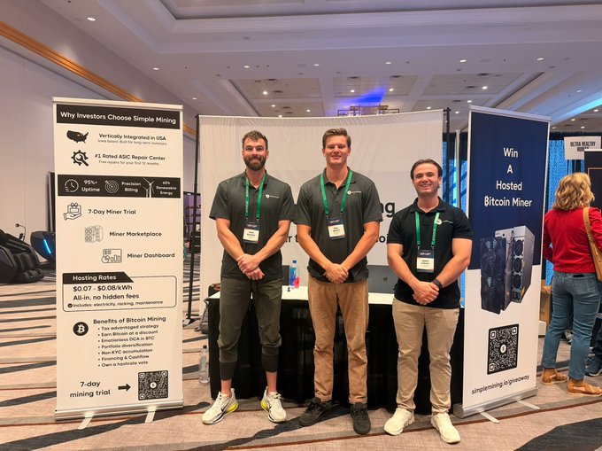 Three Simple Mining representatives pose at their booth at a finance conference, standing between banners advertising "Why Investors Choose Simple Mining" and "Win A Hosted Bitcoin Miner," illustrating the company's effort to explain Bitcoin mining to traditional finance attendees using relatable analogies like gold mining rather than technical jargon.
