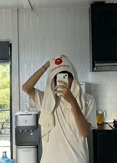 A person taking a mirror selfie in a kitchen, holding a phone and with a white towel bearing the red 'Orbiter Finance' logo draped over their head.