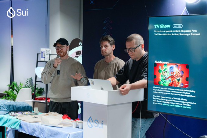 Three men presenting at a Sui-branded event, standing at a podium with a laptop, alongside a screen displaying a TV Show pitch called 'Redeowan'.