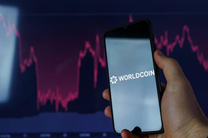 The image features a person holding up a smartphone displaying the cryptocurrency symbol "WLD" at the bottom center of the screen. In the background, there's a monitor or television screen with data related to Bitcoin and AI narratives surrounding it. The scene suggests that someone might be researching, learning about, or discussing worldcoin in relation to broader technological developments and trends in AI, such as those involving SpaceX.